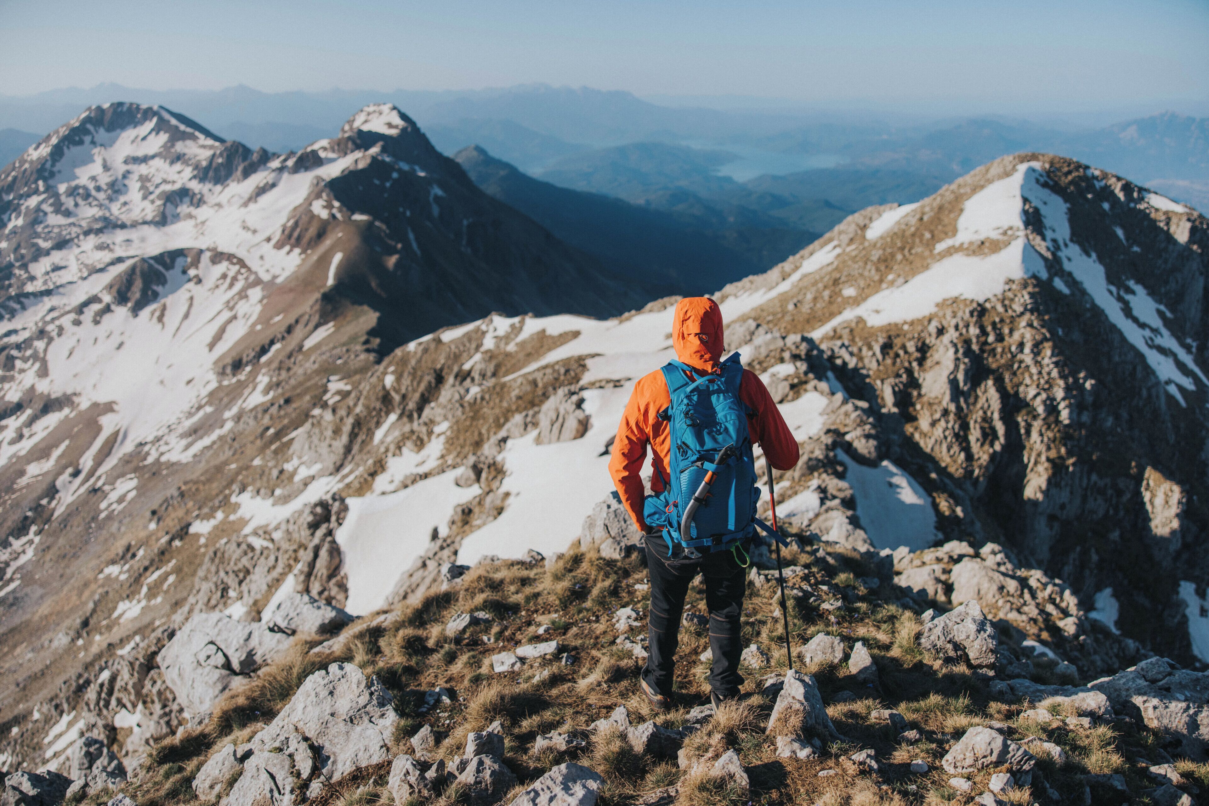 A person standing on a rocky mountain peak with a backpack, surrounded by snow-covered terrain and distant mountain ranges.
