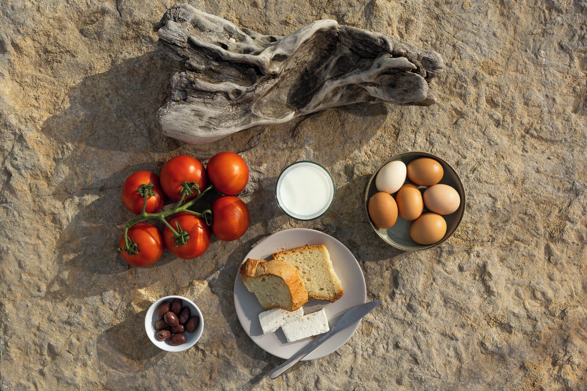 Un repas composé de tomates, d’œufs, de pain et d’olives, servi sur une surface sablonneuse.