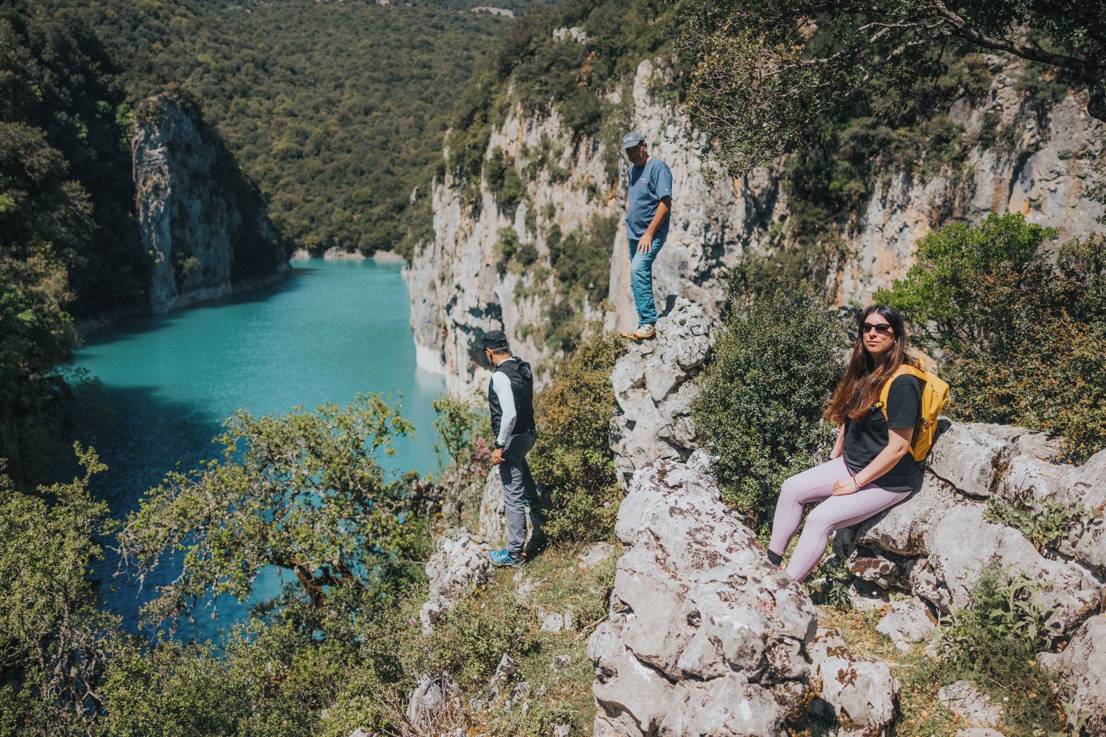 A rocky cliff with a view of a turquoise lake, a person sitting on the rocks, and another person standing on the cliff edge.