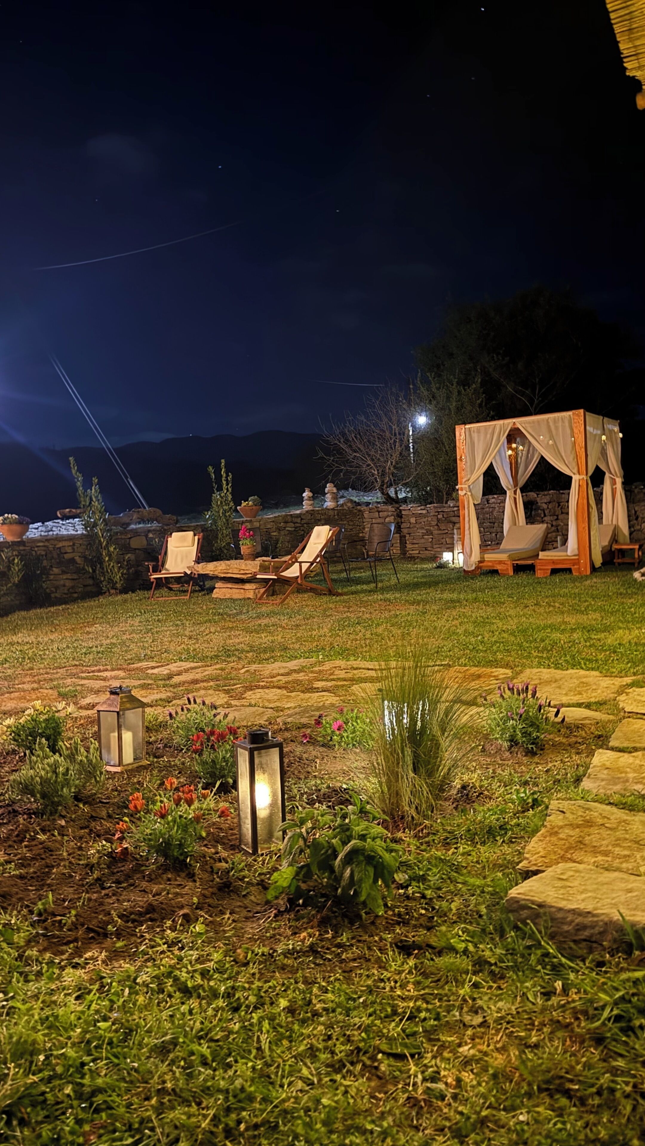 A nighttime outdoor setting with stone walls, lounge chairs, and a canopy structure.