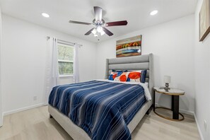 Ein Schlafzimmer mit einem Bett, einem Deckenventilator, einem Gemälde, einem Nachttisch und einem Fenster mit Jalousien.