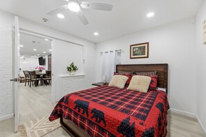 A bedroom with a bed, a ceiling fan, a window with curtains, and a dining area visible through an open door. - 3BR Near AT&T Stadium | Free Parking (Arlington)
