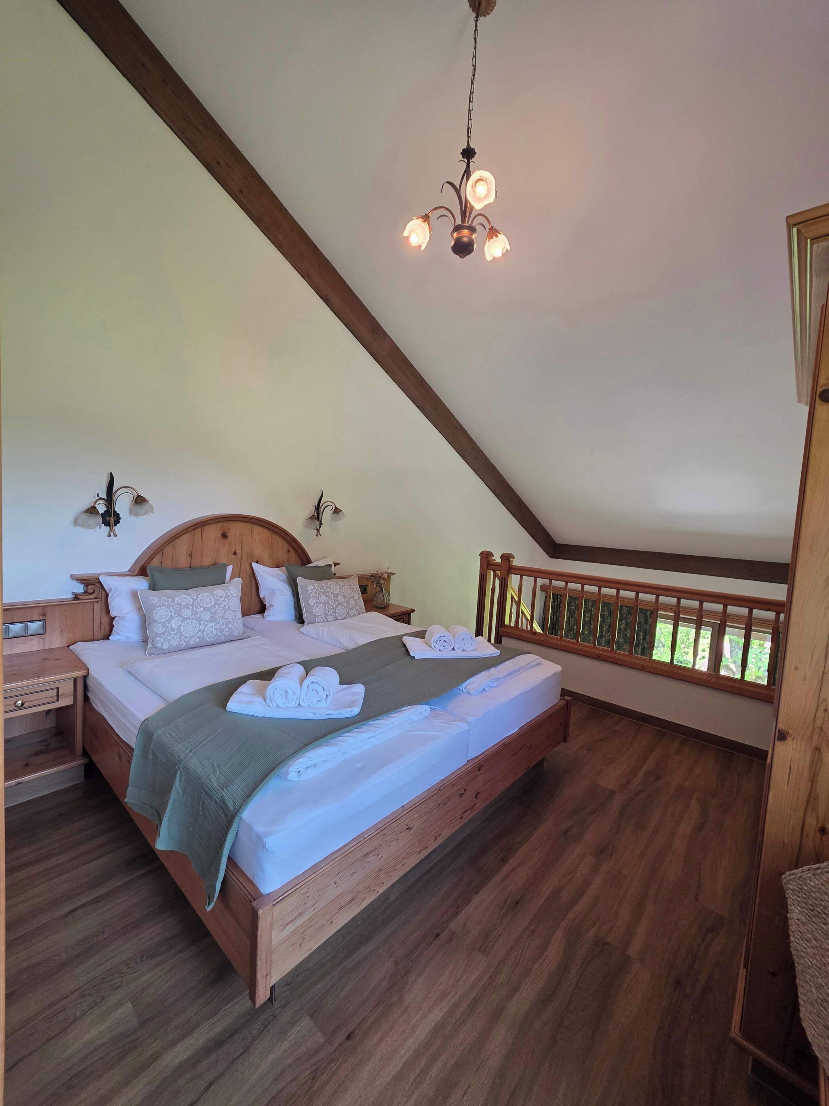 A bedroom with a wooden bed, a nightstand, a hanging light fixture, and a view of greenery through a window.