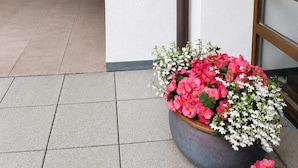 A building exterior with a white wall, a window with a number '0', a flower pot with pink and white flowers, and a black pipe. - Alpine Retreat Apt: Scenic Bavarian Nature, (Fischbachau)
