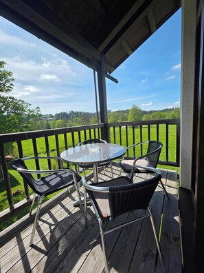 Um deck de madeira com mesa e cadeiras de metal, com vista para uma paisagem verde.