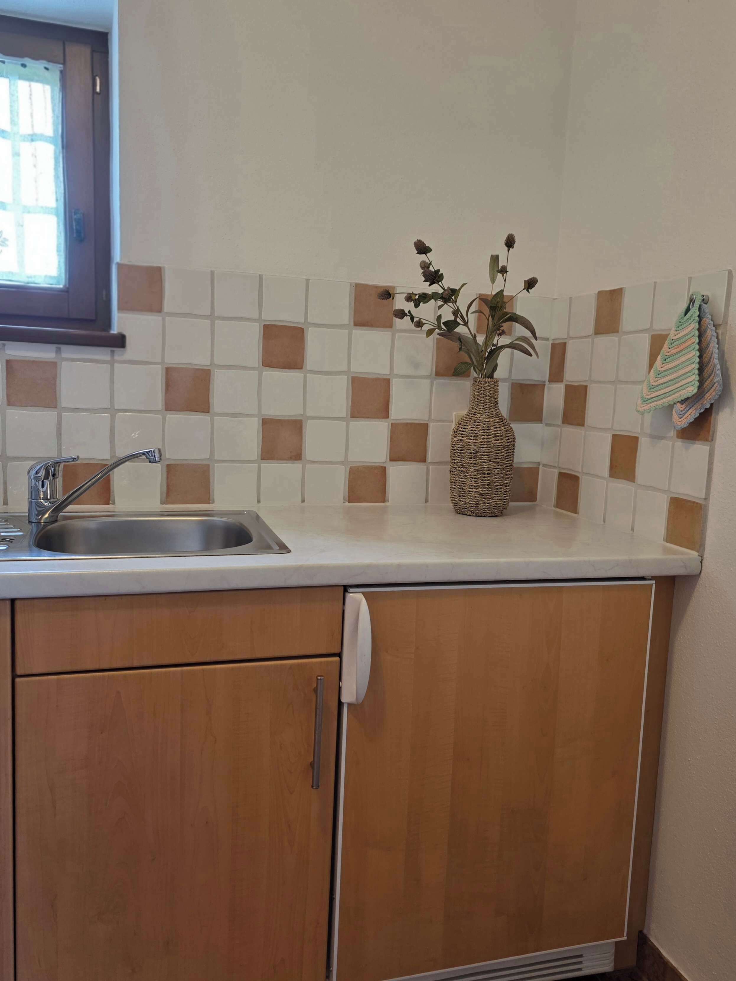 A kitchen with a sink, wooden cabinets, a tiled backsplash, and a woven vase with flowers.