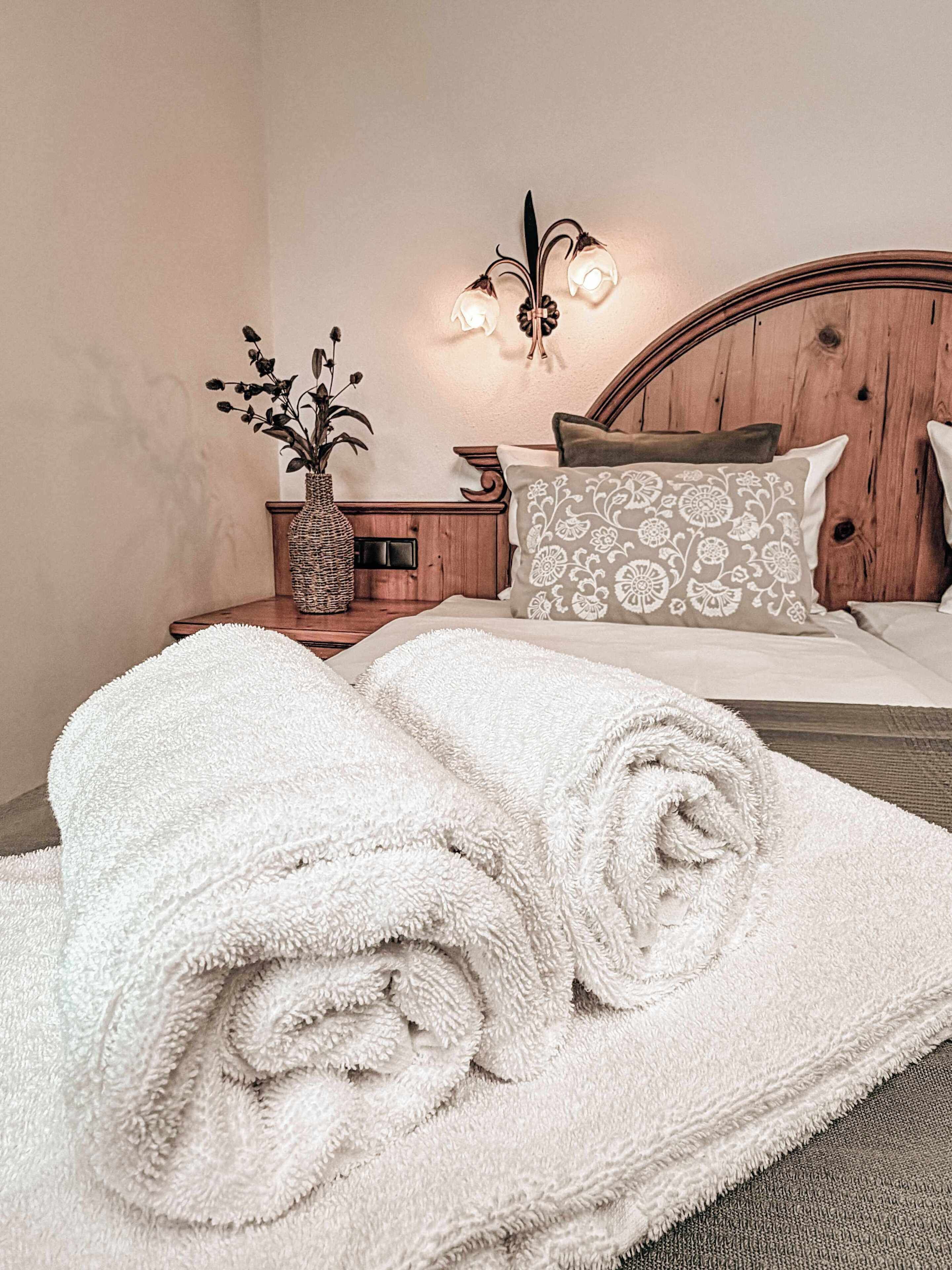 Een netjes opgemaakt bed met opgerolde witte handdoeken, een houten hoofdbord, een nachtkastje met een bloemenvaas en een wandlamp.