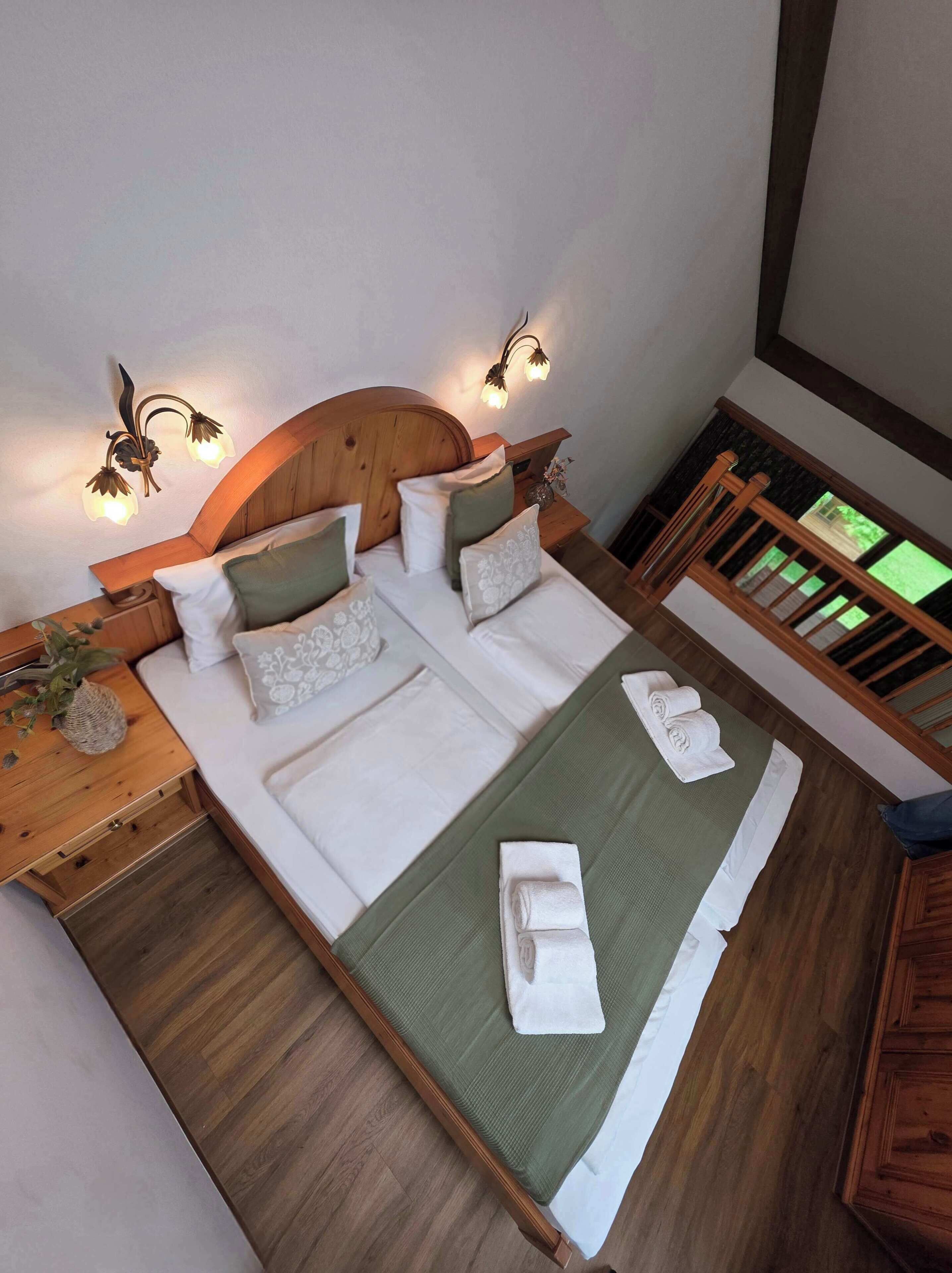 Ein Schlafzimmer mit einem Holz-Kopfteil, zwei Nachttischen mit Lampen, einem Bett mit grüner Bettwäsche und Blick ins Freie durch ein Fenster.