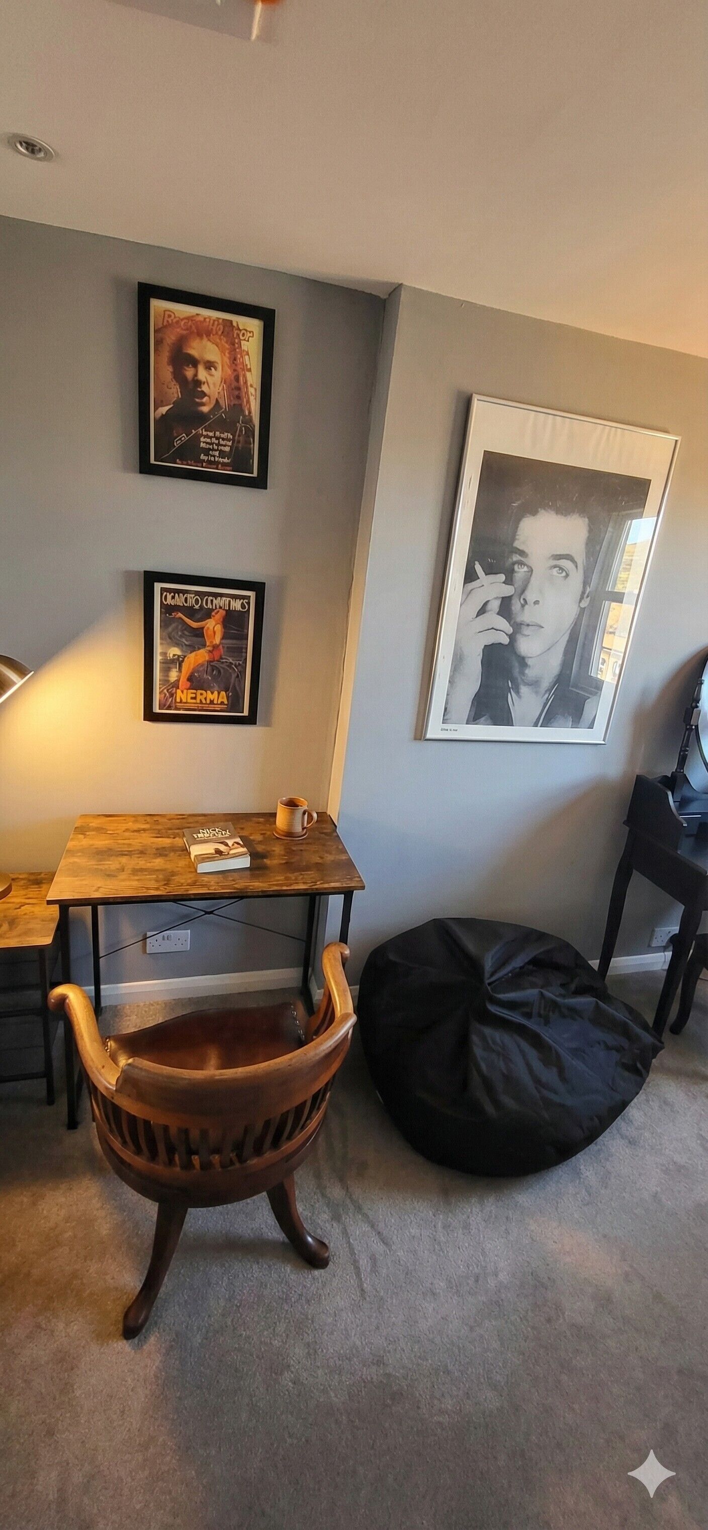 A room with a wooden chair, a black bean bag, a small table with a book and a mug, and two framed pictures on the wall.