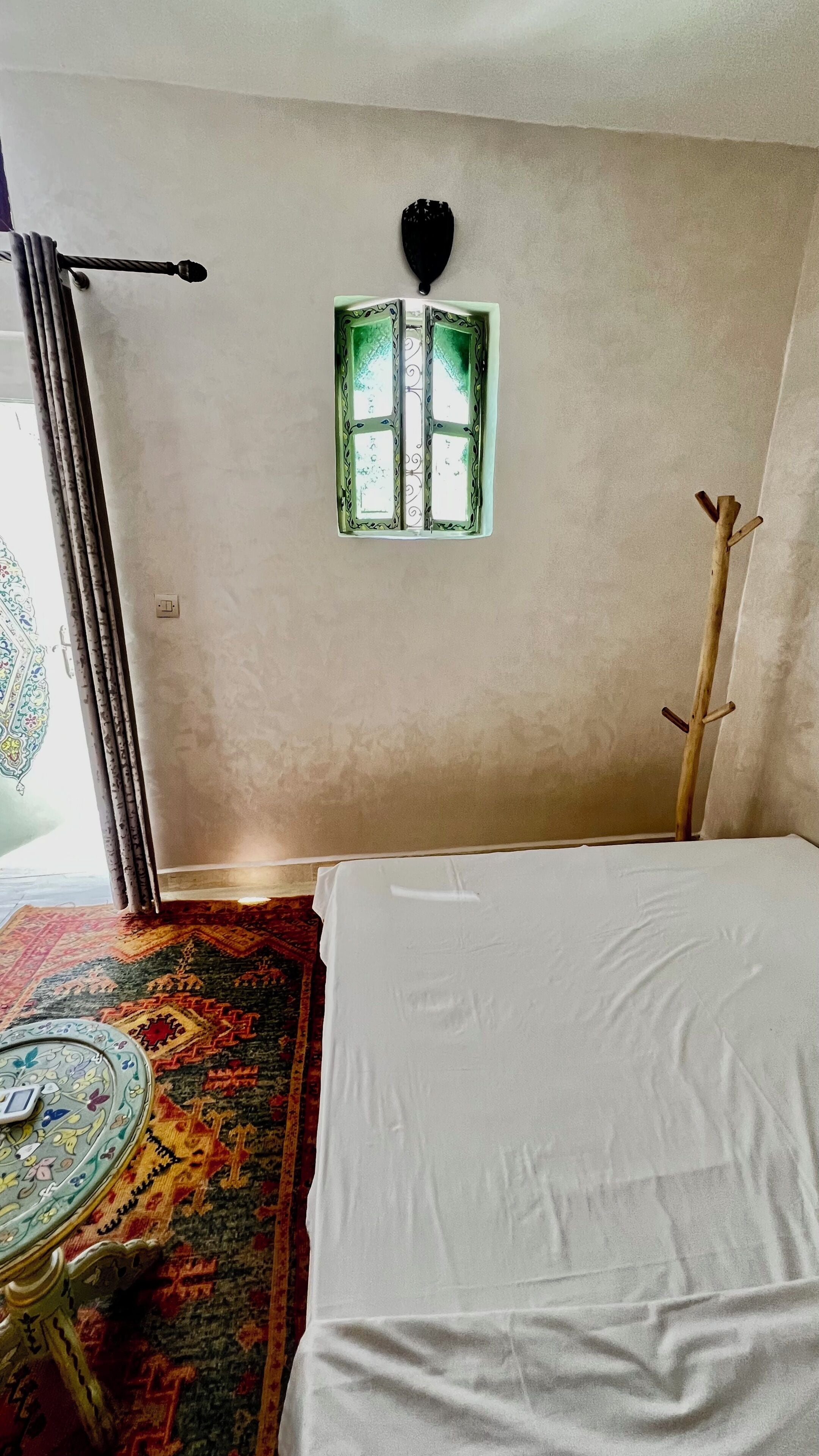 A room with a bed, a window with green curtains, a patterned rug, and a wall-mounted light fixture.