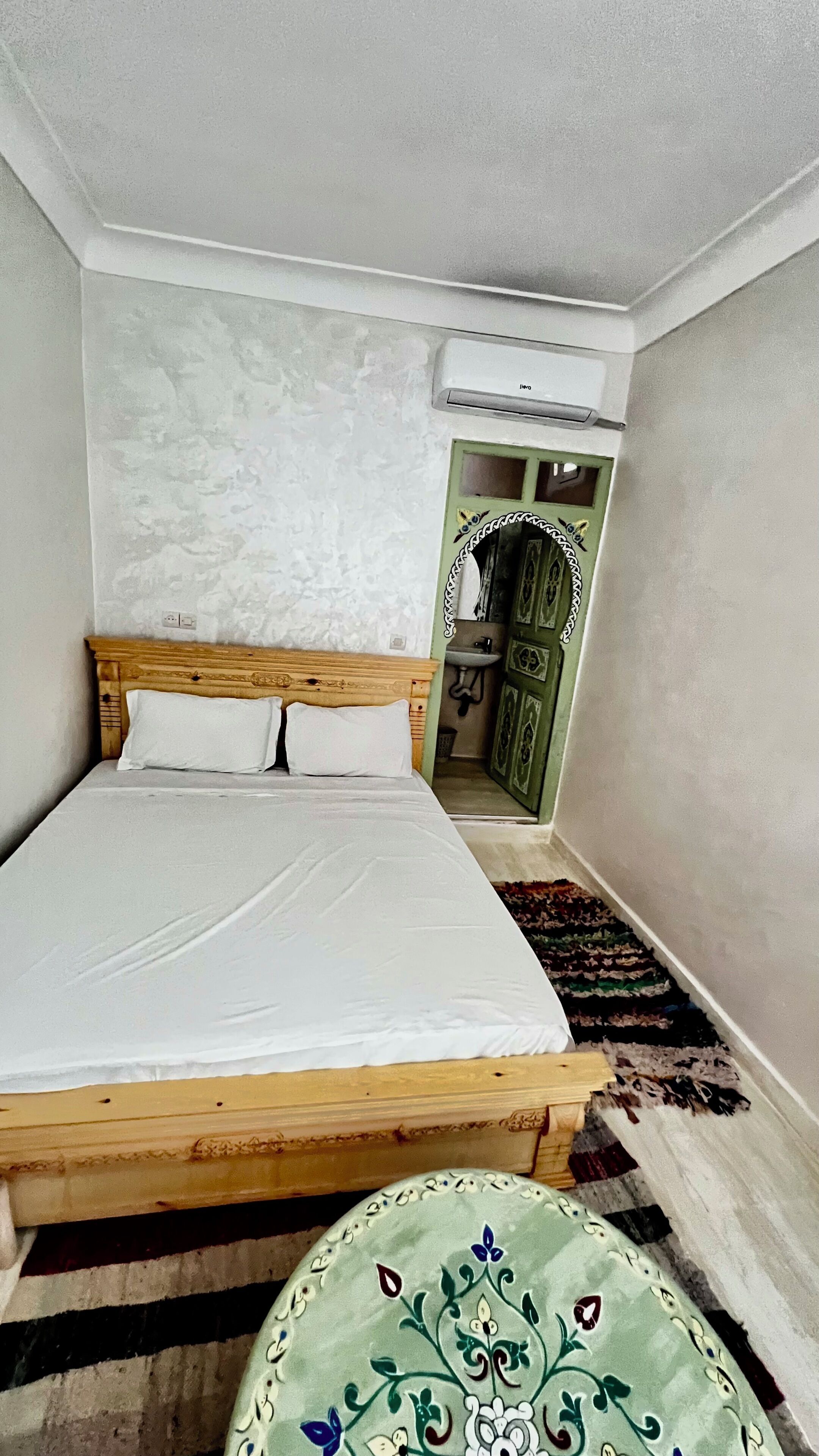 A small, narrow room with a single bed, a wooden headboard, a green door, and a small window.