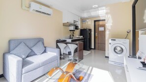 Una sala de estar moderna con sofá, una mesa de centro de vidrio, lavadora y una zona de cocina con microondas y refrigerador.