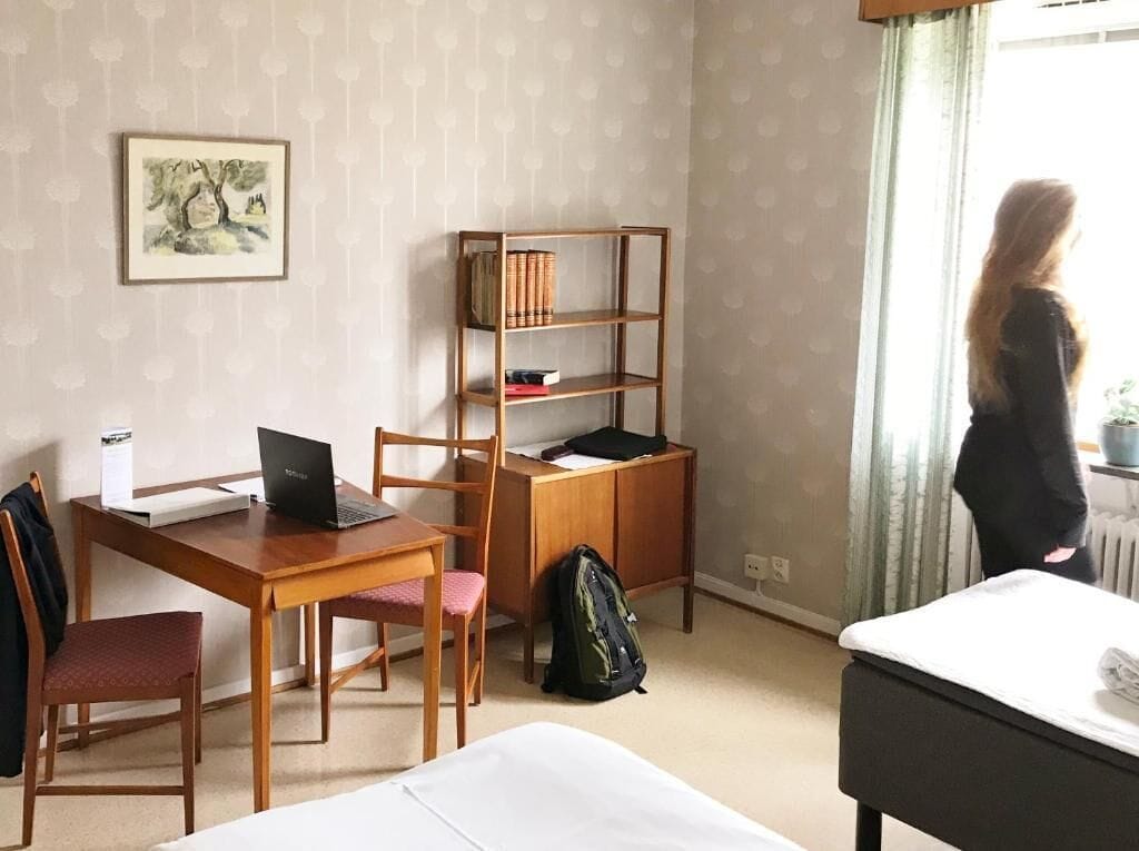 Habitación de hotel con escritorio de madera, una computadora portátil, estantería, cama y ventana con cortinas.