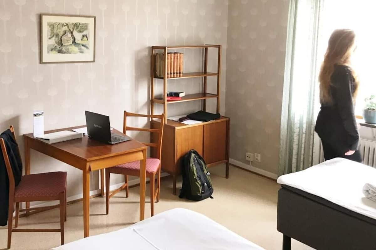 Ein Hotelzimmer mit Holzschreibtisch, einem Laptop, einem Bücherregal, einem Bett und einem Fenster mit Vorhängen.
