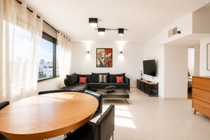 A modern living room with a sofa, a coffee table, a TV, and a dining area. - MAMAD Duplex & Panoramic Terrace near Beach | FeelHome (Tel Aviv - Jaffa)