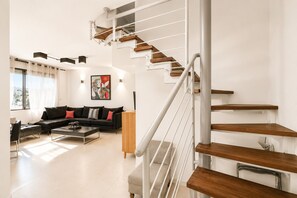 A modern living room with a staircase, a black sofa, a coffee table, and a painting on the wall. - MAMAD Duplex & Panoramic Terrace near Beach | FeelHome (Tel Aviv - Jaffa)