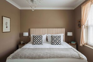 Ein ordentlich eingerichtetes Schlafzimmer mit einem großen Bett, zwei Nachttischen, einer Kandelaberleuchte und einem Fenster mit Vorhängen.