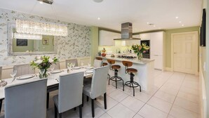 A modern kitchen with white cabinets, a central island with bar stools, a dining area with a chandelier, and a mirrored wall. - The Monmouth House of Bath (Bath)