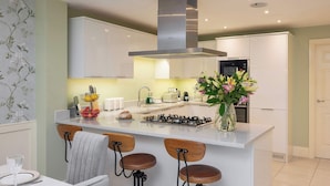 A modern kitchen with white cabinets, a stainless steel range hood, a central island with stools, and a vase of flowers. - The Monmouth House of Bath (Bath)