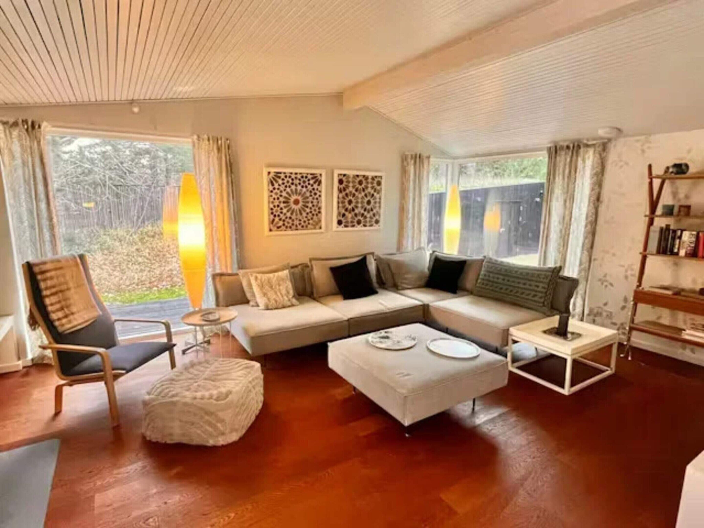 A living room with a sectional sofa, a small round table, a white ottoman, and a bookshelf.