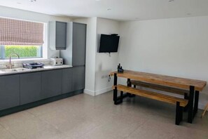 A modern kitchen with grey cabinets, a wooden bench, a TV mounted on the wall, and a window with a checkered blind. - Coome Lodge: Modern Stay near Saunton Sands Beach (Braunton)