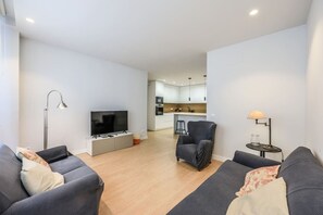Una sala moderna con un televisor de pantalla plana, dos sofás, una mesa de centro, una lámpara de pie y una cocina al fondo.