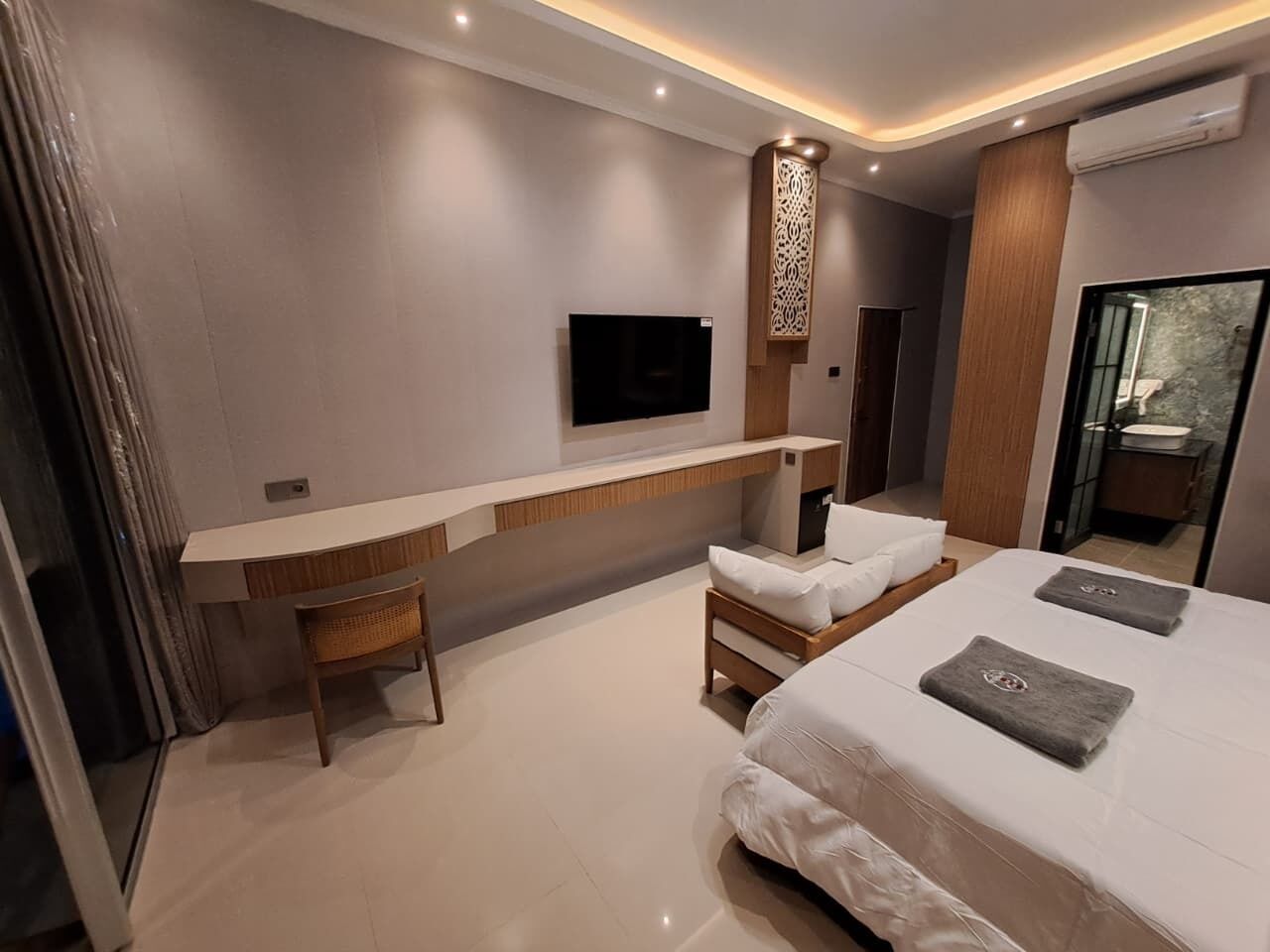 A modern hotel room with a bed, a desk, a chair, a TV, and a bathroom visible through a glass door.