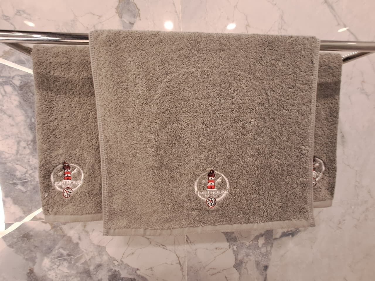 Three towels with a lighthouse logo on a metal towel rack against a marble wall.