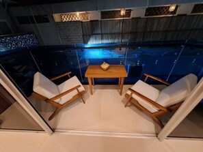 A balcony with a glass partition, two white cushioned chairs, a wooden table, and a blue-lit background. - Lighthouse Hotel (Gili Trawangan)