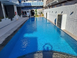 Una piscina con superficie de agua azul cristalina, rodeada por un edificio moderno con fachada estampada y un muro de piedra.