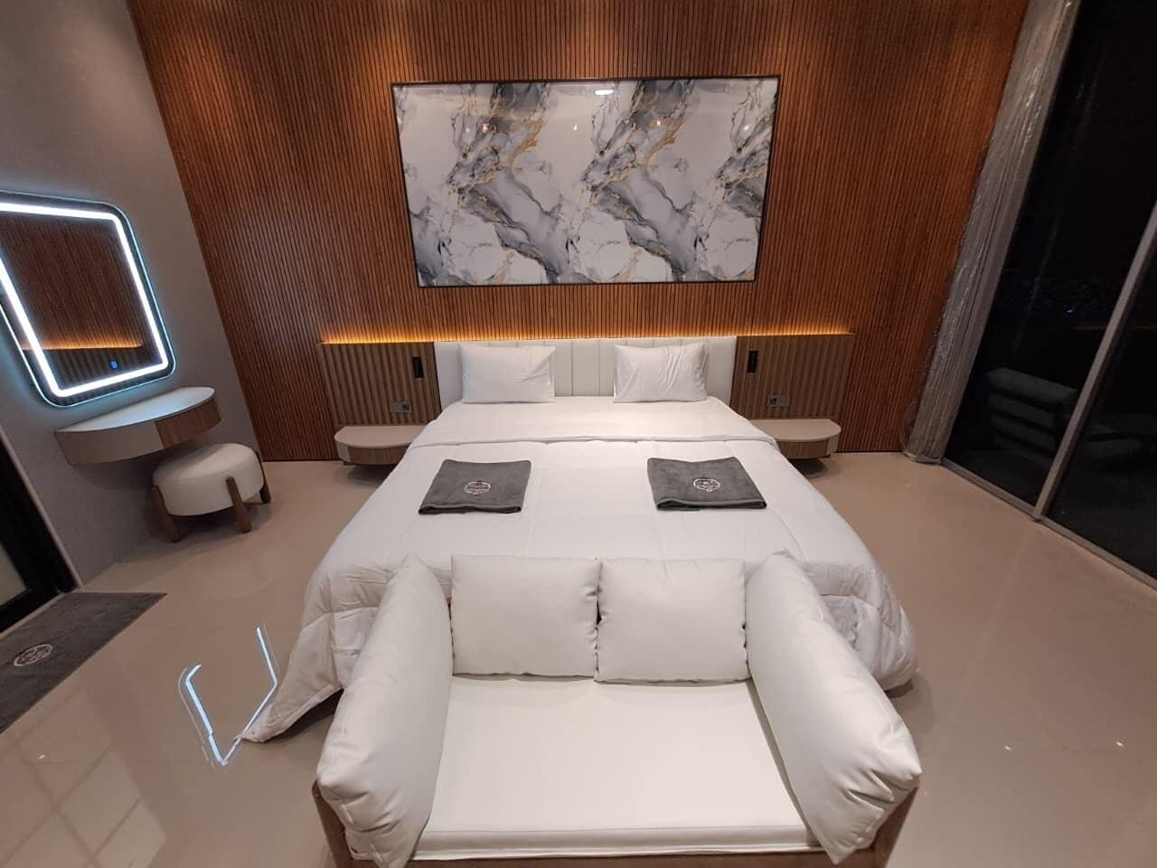 A modern bedroom with a large bed, a seating area, a TV mounted on the wall, and a wooden panel design.