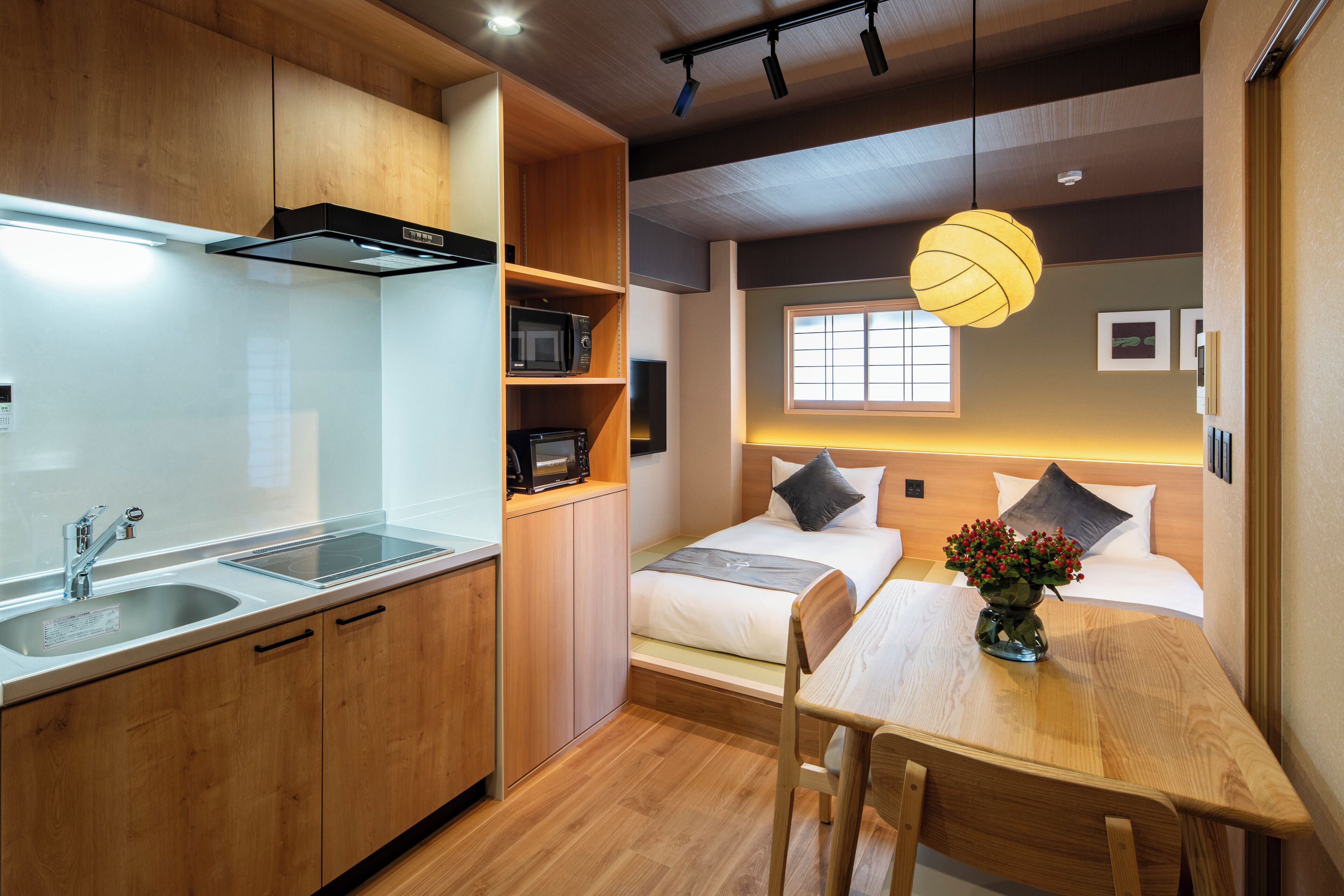 A compact hotel room with a kitchenette, a bed with pillows, a bedside table with a vase, and a window.