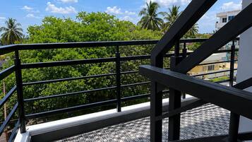 Un balcon doté d’une rambarde en métal, d’un sol carrelé et offrant une vue sur la verdure et les bâtiments, le tout sous un ciel dégagé.