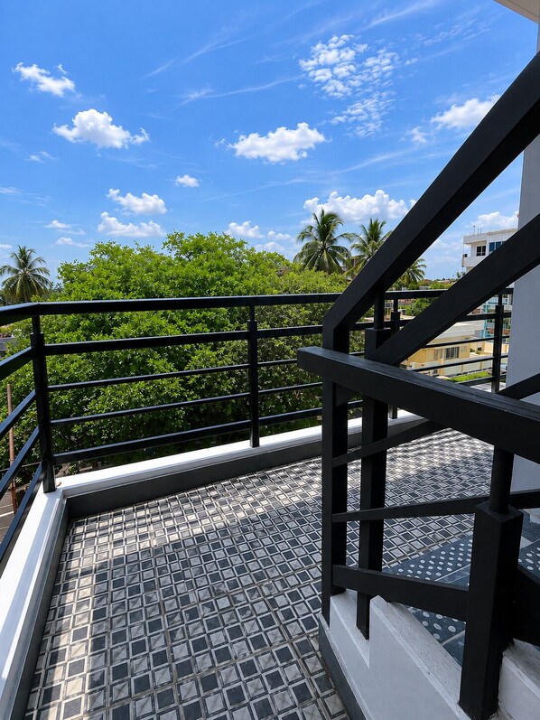 A balcony with a metal railing, a tiled floor, and a view of greenery and buildings under a clear sky.