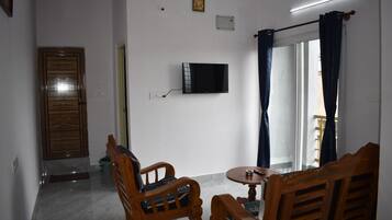 Une pièce comprenant une télévision, deux chaises en bois, une petite table, une porte et une fenêtre avec des rideaux.