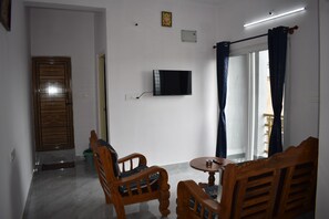 A room with a television, two wooden chairs, a small table, a door, and a window with curtains.