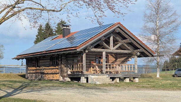 Una cabaña de troncos con techo inclinado, paneles solares y un porche con asientos.