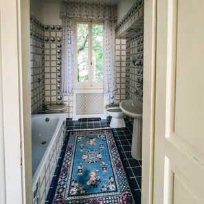 Et aflangt badeværelse med badekar, toilet og vask, beklædt med fliser i et gentaget mønster, samt et vindue og en tæppe med et mønster.