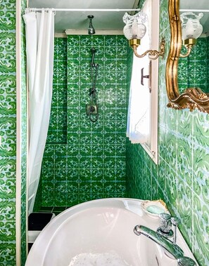 A bathroom with green patterned tiles, a white bathtub, a shower with a gold faucet, and a window with a white curtain.