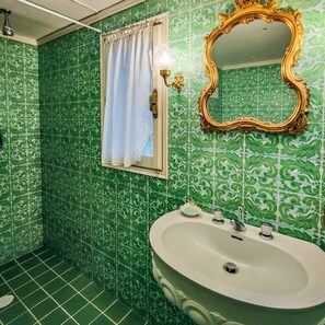 Une salle de bain avec des carreaux verts Ă motifs, un lavabo blanc, un miroir avec un cadre dorĂ© et une fenĂȘtre ornĂ©e dâun rideau blanc.