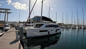 En lystbådehavn med flere sejlbåde i havnen, en træbro og en moderne bygning i baggrunden.