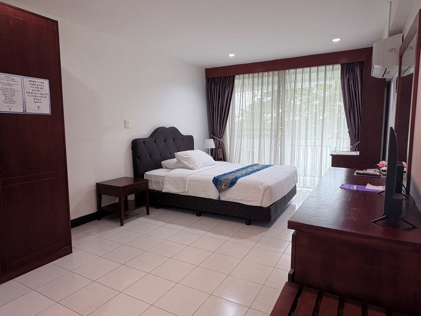 Quarto de hotel com cama, mesa de cabeceira, escrivaninha e janela com cortinas.