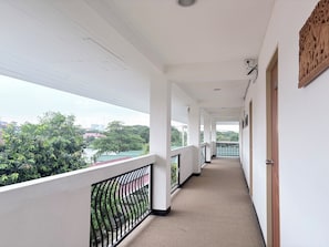 Un pasillo con vistas a zonas verdes y edificios, paredes blancas y barandilla negra.
