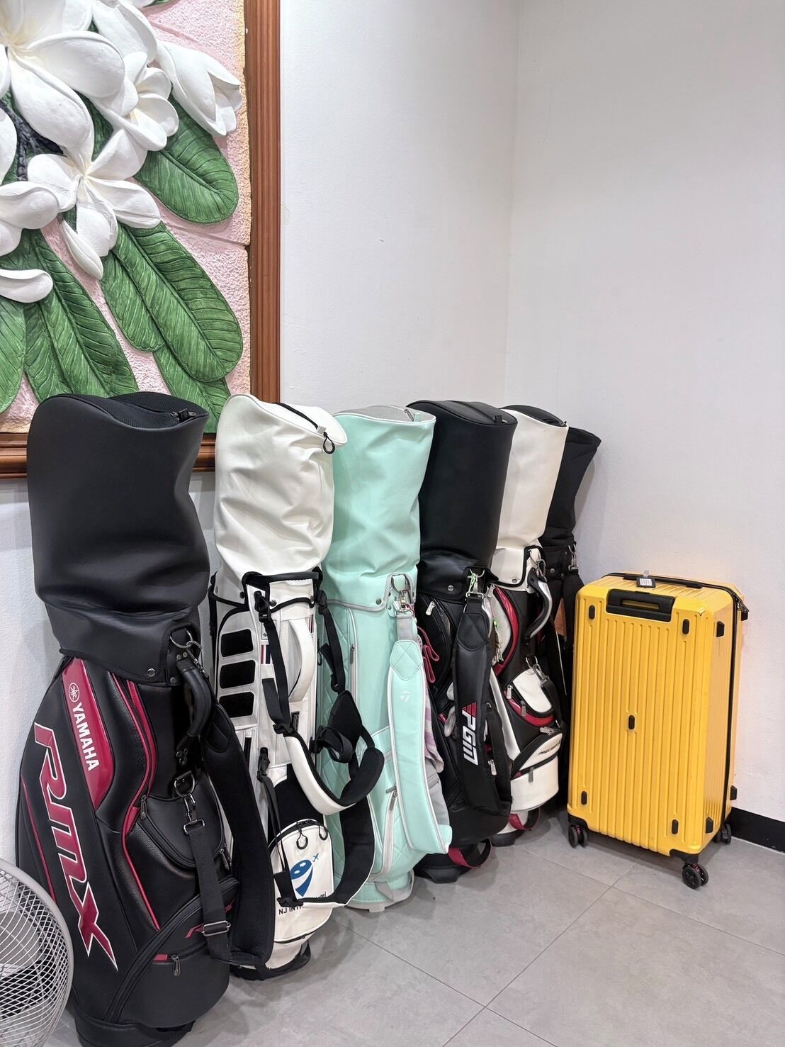 A corner of a room with a wall-mounted fan, a window with a floral decoration, and a row of luggage including a black Yamaha backpack, a white and teal backpack, a black and white backpack, and a yellow suitcase.