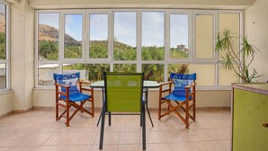 Un balcón con mesa y sillas, ventanas amplias y vista a un paisaje montañoso.