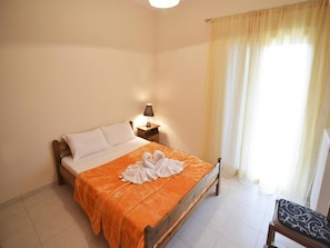 Un dormitorio con cama, mesita de noche con lámpara, una silla y una ventana con cortinas.
