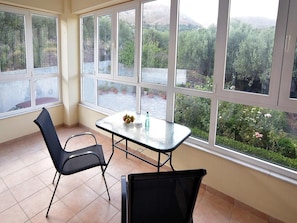 Quarto com mesa e cadeiras de vidro, grandes janelas com vista para vegetação e colinas.