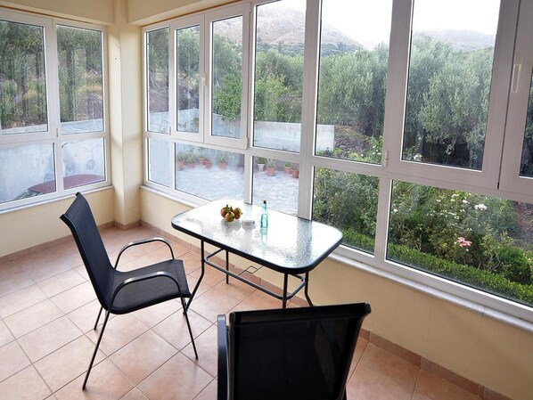 Une pièce avec une table et des chaises en verre, de grandes fenêtres donnant sur un paysage verdoyant et des collines.