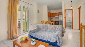 Un espacio habitable compacto con sofá, una mesa de comedor con frutero, una cocina con armarios y electrodomésticos, y un ventanal corredizo que da a un área exterior.