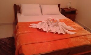 A neatly made bed with white pillows and an orange bedspread, a wooden nightstand with a vase, and a small rug.
